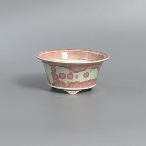 May include: A ceramic bowl with a wide, flared rim and three small feet. The exterior features a pale green base glaze with mottled, circular patches of dusty rose and mauve. The interior rim is finished in a soft, muted pink tone.