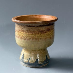 May include: A ceramic chalice-shaped vessel featuring a wide, flared rim and a pedestal base. The exterior displays a gradient of earthy tones, transitioning from a warm terracotta brown at the top to a mottled yellow-green and a dark charcoal base.
