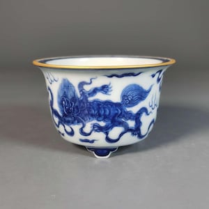 Puede incluir: Un cuenco de cerámica blanco y azul con borde dorado. El cuenco presenta un detallado diseño de dragón azul. El cuenco tiene tres pequeños pies y un borde acampanado. El diseño es un motivo asiático tradicional.