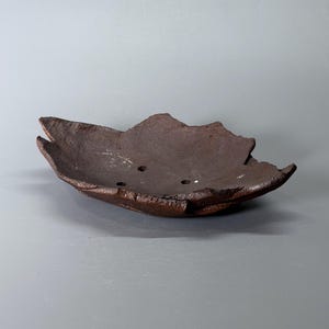 May include: A dark brown, shallow ceramic bowl with a rough, textured surface and an irregular, leaf-like edge. The interior of the bowl features three small, circular drainage holes evenly spaced across the bottom, creating a functional design.