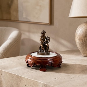 Puede incluir: Una escultura de bronce de una figura sentada tocando una pequeña lira, montada sobre una base circular de mármol blanco. Esta base se asienta sobre un soporte de madera marrón oscuro con patas talladas y volutas decorativas.
