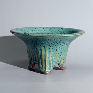 Pode incluir: Uma taça de cerâmica turquesa com uma borda larga e alargada e um padrão salpicado de azul escuro. A taça apresenta detalhes nervurados verticais ao longo do corpo exterior e assenta em três pequenos pés curvos de argila castanho-avermelhada.