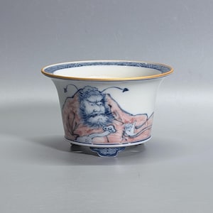 May include: A white ceramic bowl with a flared rim and three small feet. The exterior features a hand-painted blue and reddish-brown illustration of a bearded figure with prominent eyebrows, accented by a blue geometric pattern along the inner rim.