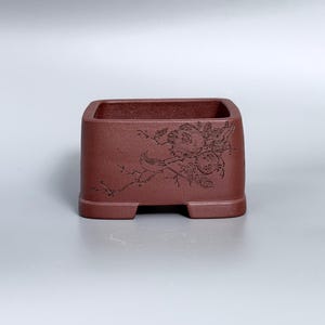 May include: A square, reddish-brown ceramic planter with a detailed floral and bird design etched on the side. The planter has a textured surface and sits on four small feet. The open top is square.
