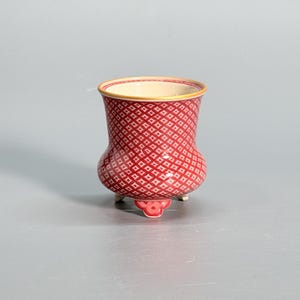 Puede incluir: Un pequeño recipiente de cerámica con un exterior rojo vibrante y un patrón de celosía de diamantes blancos repetido. El contenedor tiene un borde acampanado, un cuerpo redondeado y se apoya sobre tres pies decorativos. El interior es crema.