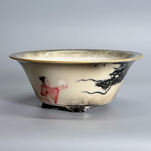 May include: A ceramic bowl with a cream-colored base and a dark, speckled gradient rim. The exterior features a black ink-style dragon illustration and a figure in a red robe holding a white orb. The bowl rests on three small, dark feet.
