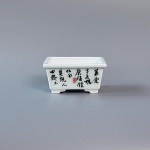 May include: A small, rectangular white ceramic planter with rounded corners and four small feet. Black calligraphy and red stamps decorate the sides. The planter is designed for bonsai or small plants.