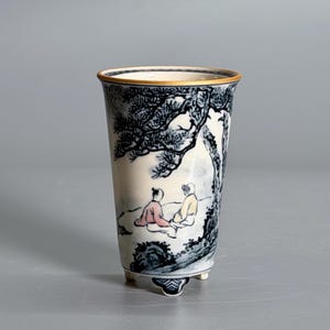May include: A tapered ceramic cup with three small feet, featuring a blue and white ink-wash style landscape painting of two figures sitting under a pine tree, with subtle pink and yellow accents on their clothing and a gold-toned rim.
