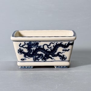 May include: A rectangular ceramic planter with an off-white base colour and a dark blue hand-painted dragon motif wrapping around the exterior. The rim and the four small feet feature a geometric blue pattern, creating a distinct contrast.