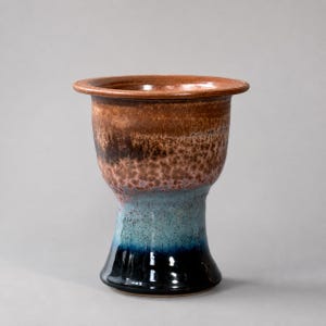 May include: A ceramic chalice featuring a wide, flared rim and a tapered base. The glaze transitions from a warm, mottled brown at the top to a vibrant turquoise in the middle, ending in a deep, glossy black at the bottom of the vessel.