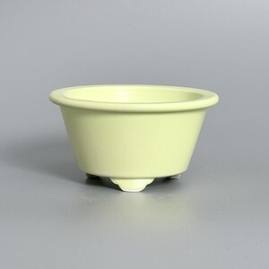 Puede incluir: Un cuenco de cerámica de color amarillo pálido con un acabado mate suave y un borde ancho y redondeado. El cuenco presenta un cuerpo cónico que se estrecha y descansa sobre tres pies pequeños y uniformes, ofreciendo una base estable.