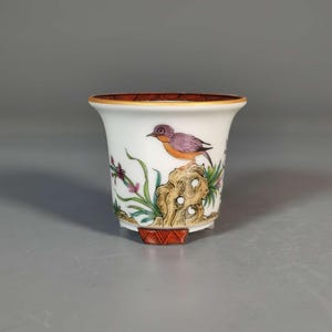 May include: A white porcelain cup with a flared rim, featuring a painted bird perched on a rock with colorful foliage. The rim has a brown, wood-grain design, and the base has a red geometric pattern. The bird has purple and orange feathers.