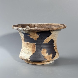 May include: A ceramic bowl with a flared rim, featuring a textured, speckled beige and tan exterior surface contrasted by bold, irregular patches of matte black glaze that wrap around the midsection of the vessel, creating a distinct visual pattern.