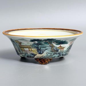 May include: A shallow ceramic bowl with a wide rim and three small feet. The exterior features a detailed landscape painting with green pine trees, rocky mountains, and small buildings in shades of yellow, blue, and green on a white base.