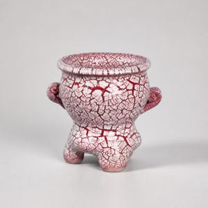 May include: A small ceramic pot featuring a rounded body with two side handles and three short legs. The exterior surface is finished with a white glaze overlaid by a deep red crackle pattern that covers the entire vessel from rim to base.