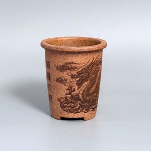 Op de afbeelding: Een cilindervormige, terracotta-kleurige keramische plantenpot met een gespikkelde afwerking. De buitenkant is voorzien van een donkerbruin gegraveerd drakenmotief tussen wolken en verticale Chinese karakters. De pot heeft een brede, ronde rand.