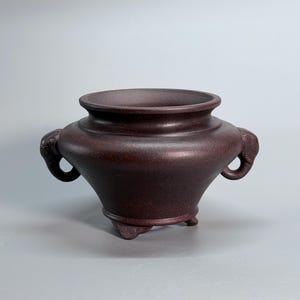 Puede incluir: Un recipiente de cerámica de color marrón rojizo oscuro con un cuerpo ancho y redondeado y un borde acampanado. La pieza presenta dos asas esculpidas en forma de cabeza de elefante en los lados opuestos y se apoya sobre tres pies pequeños.