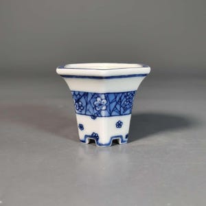 Op de afbeelding: Een kleine, vierkante keramische plantenpot met een witte basis en blauwe handgeschilderde details. Het ontwerp bevat een band met ingewikkelde geometrische patronen afgewisseld met kleine bloemmotieven, rustend op vier korte, stevige voetjes.