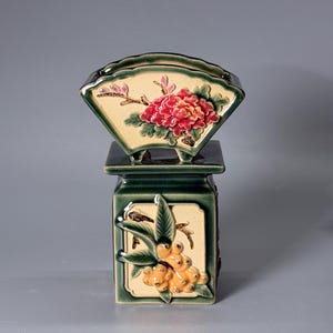 May include: A ceramic vase featuring a square base with a relief of yellow berries and green leaves on a cream panel, topped with a fan-shaped section displaying a relief of a large red flower and pink buds on a cream background, all in dark green.