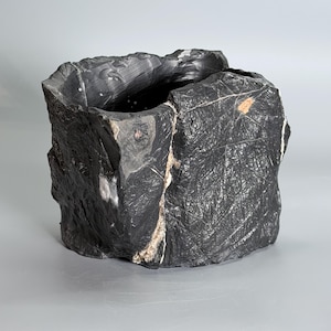 May include: A dark charcoal gray stone vessel with a rough, craggy exterior texture. The surface features intricate white veining and subtle tan mineral inclusions, with a wide, hollow opening at the top and jagged, uneven edges.