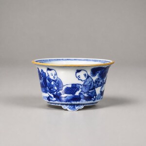 May include: A small porcelain bowl featuring blue and white underglaze hand-painted illustrations of two children playing a board game outdoors. The bowl has a flared rim with a thin gold-toned edge and rests on three small, shaped feet.