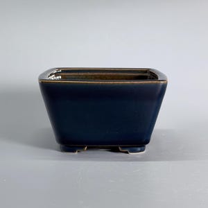 Puede incluir: Una maceta cuadrada de cerámica con un acabado esmaltado en azul marino profundo y un borde marrón en contraste. El recipiente presenta una silueta cónica con cuatro pequeños pies integrados en la base, proporcionando una base estable.