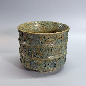 May include: A cylindrical ceramic bowl featuring a heavily textured, porous surface with a mottled finish in shades of dark green, bronze, and brown. The exterior wall displays three horizontal ridges, creating a layered, rugged appearance.