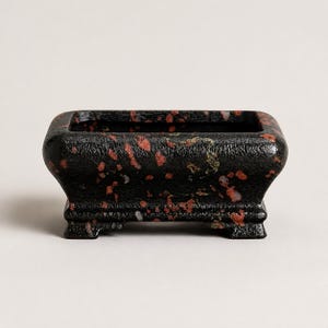 May include: A rectangular planter with a textured black finish and scattered red and orange speckles. The vessel features a wide opening, rounded corners, and a raised base with four small feet, providing a stable foundation for small plants.