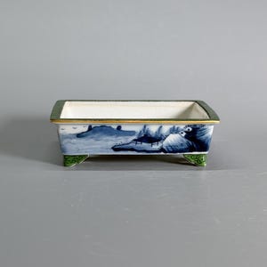 Puede incluir: Una maceta rectangular de cerámica para bonsái con interior blanco y una ilustración de paisaje azul en el exterior. El borde está acentuado con una fina línea dorada y un borde verde oscuro, mientras que los cuatro pies están pintados de verde claro.