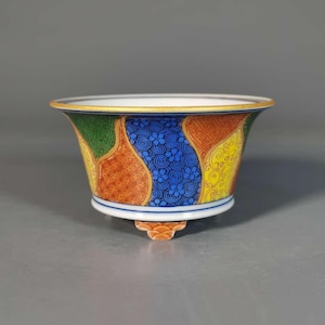 May include: A small, decorative ceramic bowl with a flared shape. The bowl has a white interior and a colourful exterior with a gold rim. The exterior is decorated with a patchwork of patterns in green, blue, orange, and yellow. The bowl rests on a small, decorative foot.