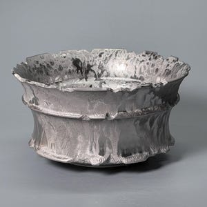 Puede incluir: Un cuenco de metal plateado con un borde ancho y acampanado y una superficie texturizada e irregular. El cuenco presenta un reborde horizontal distintivo alrededor de su centro y un acabado moteado en gris oscuro y plata con un patrón complejo.