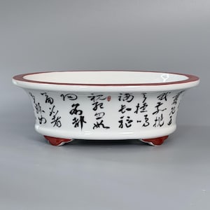 May include: A white ceramic oval bonsai pot featuring a dark red rim and base. The exterior surface is decorated with black Chinese calligraphy characters. The vessel has a wide, open top and is supported by three small, integrated feet.