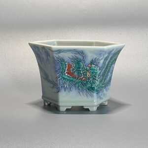 Może przedstawiać: Jasnoniebieska, sześciokątna ceramiczna donica z szczegółową ilustracją smoka w odcieniach zieleni, czerwieni i niebieskiego. Donica ma lekko rozszerzający się kształt i stoi na czterech małych nóżkach. Wzór smoka znajduje się z przodu.