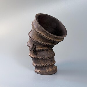 May include: A dark brown, cylindrical ceramic vessel with a distinct, angled tilt. The exterior surface features four horizontal, raised bands decorated with a repeating pattern of small, circular beads or dots, creating a textured appearance.