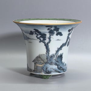 May include: A porcelain planter featuring a hand-painted landscape scene with blue pine trees, a small hut, and mountains. The rim and base are decorated with a green patterned border, and the top edge is finished with a thin gold-colored trim.