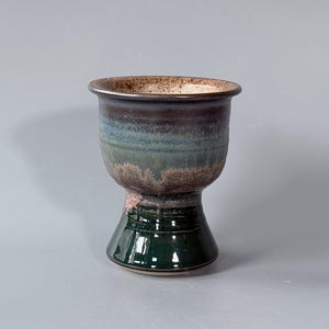 May include: A ceramic goblet with a wide, flared rim and a tapered base. The upper portion features a gradient of blue, green, and brown tones with a textured, speckled finish. The lower section is a solid, glossy dark green with a smooth surface.