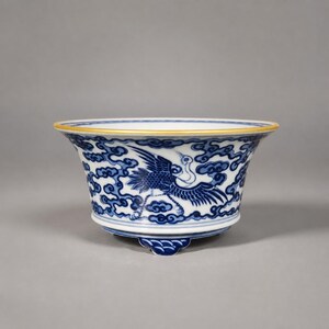 May include: A porcelain bowl featuring a white base with intricate blue underglaze patterns of flying cranes and stylized clouds. The bowl has a flared rim with a thin yellow border and rests on a small, scalloped base with blue wave motifs.