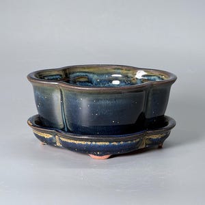 Puede incluir: Una maceta de cerámica para bonsái con un borde festoneado en forma de flor y un plato a juego. La pieza presenta un esmalte reactivo azul profundo y verde oscuro con sutiles motas doradas. La maceta descansa sobre pies pequeños y cortos.
