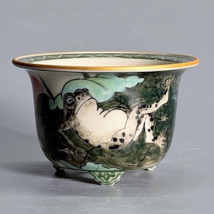 May include: A ceramic planter with a flared rim and three small feet. The exterior features a hand-painted illustration of a spotted frog resting on a large green leaf. The rim is accented with a thin gold band and a green geometric pattern.