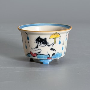 May include: A small ceramic bowl with a flared rim featuring a hand-painted illustration of a black and white cat holding a yellow umbrella. The cat wears red boots and stands on a blue puddle amidst falling blue raindrops.