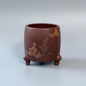 May include: A dark brown cylindrical vessel with a textured surface, supported by three small rounded feet. The exterior features a raised, light-colored relief design depicting a cluster of small flowers and foliage on the front side.