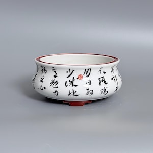 May include: A white ceramic bowl with a rounded shape and a dark red rim. The exterior features black Chinese calligraphy characters and a small red seal stamp. The base of the bowl is also dark red, providing a contrast to the white body.