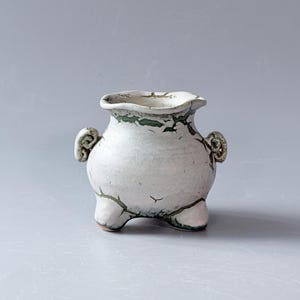 Può includere: Un piccolo vaso in ceramica color bianco sporco con un bordo largo e ondulato e due manici laterali arricciati. La superficie presenta un distinto motivo a smalto craquelé con linee verde scuro che accentuano le crepe, su tre piedi corti.