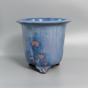 May include: A ceramic planter with a flared rim and a glossy blue glaze featuring mottled pinkish-brown patches. The vessel is supported by three small, ornate feet at the base, showcasing a smooth, reflective surface and a wide, open top.
