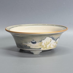 May include: A shallow ceramic bowl with a wide, flared rim featuring a thin gold-toned edge. The exterior is decorated with a hand-painted landscape scene in muted blue, green, and cream tones, depicting a small structure and foliage.