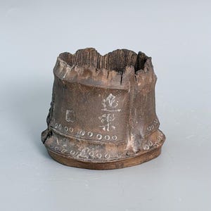 May include: A dark brown, weathered wooden vessel with a rough, uneven top edge. The exterior features a series of small, circular indentations along the base and two distinct white characters etched into the side, resembling a rustic, organic container.