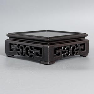 May include: A square dark brown wooden display stand featuring a flat top surface and an openwork apron with intricate geometric patterns on all four sides, supported by four short, sturdy feet at the corners of the base structure.