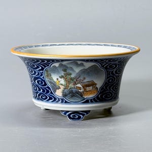 Puede incluir: Un cuenco de cerámica con borde acampanado y tres pies pequeños. El exterior presenta un patrón de remolinos azul oscuro que rodea una ilustración paisajística central de una casa, árboles y una figura en amarillo. El borde tiene una fina línea dorada.
