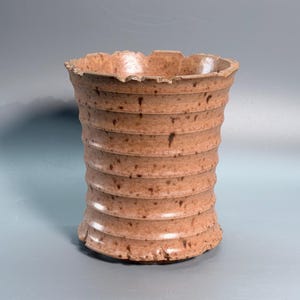 May include: A cylindrical ceramic vase with a light brown, speckled finish and a series of horizontal ridges running along the exterior. The top and bottom edges feature a jagged, uneven, and textured rim design, giving the vessel a distinct appearance.