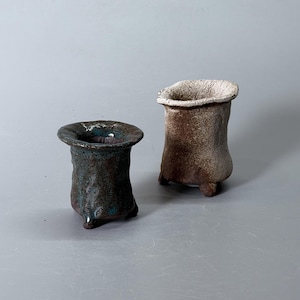 May include: Two small, hand-formed ceramic vessels with flared rims and three short feet. The left vessel features a dark, mottled blue and brown glaze, while the right vessel has a textured, light beige and brown finish with a rough, porous rim.